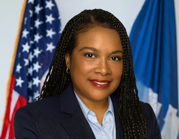 Portrait of Jhané Wilcox, Director of Communications, smiling in a navy suit with the American flag visible in the background.